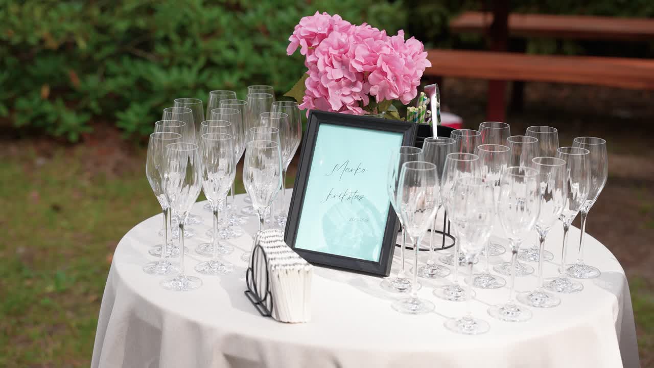 Elegante estación de bebidas al aire libre con copas de champán transparentes, flores de hortensia rosas, servilletas y un cartel con marco, lista para una celebración o evento especial.