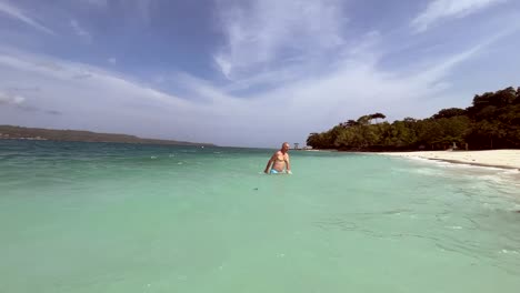 Ein-Mann-Schwimmt-In-Einem-Blauen,-Klaren-Wasser-Mit-Einem-Weißen-Sandstrand-Und-Einem-Blauen-Himmel