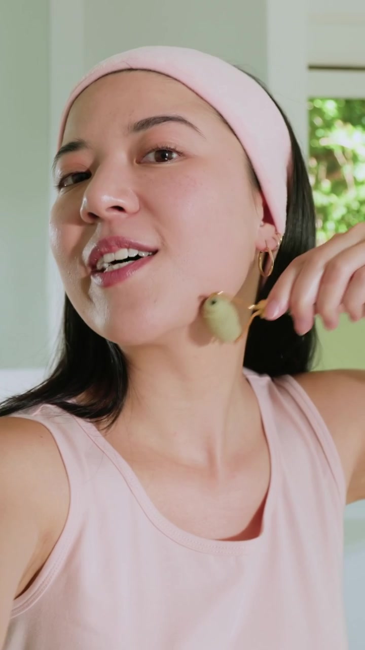 Video vertical: Mujer con diadema rosa hablando a la cámara, usando un rodillo verde para la piel en el lavabo, bloqueando la lente.