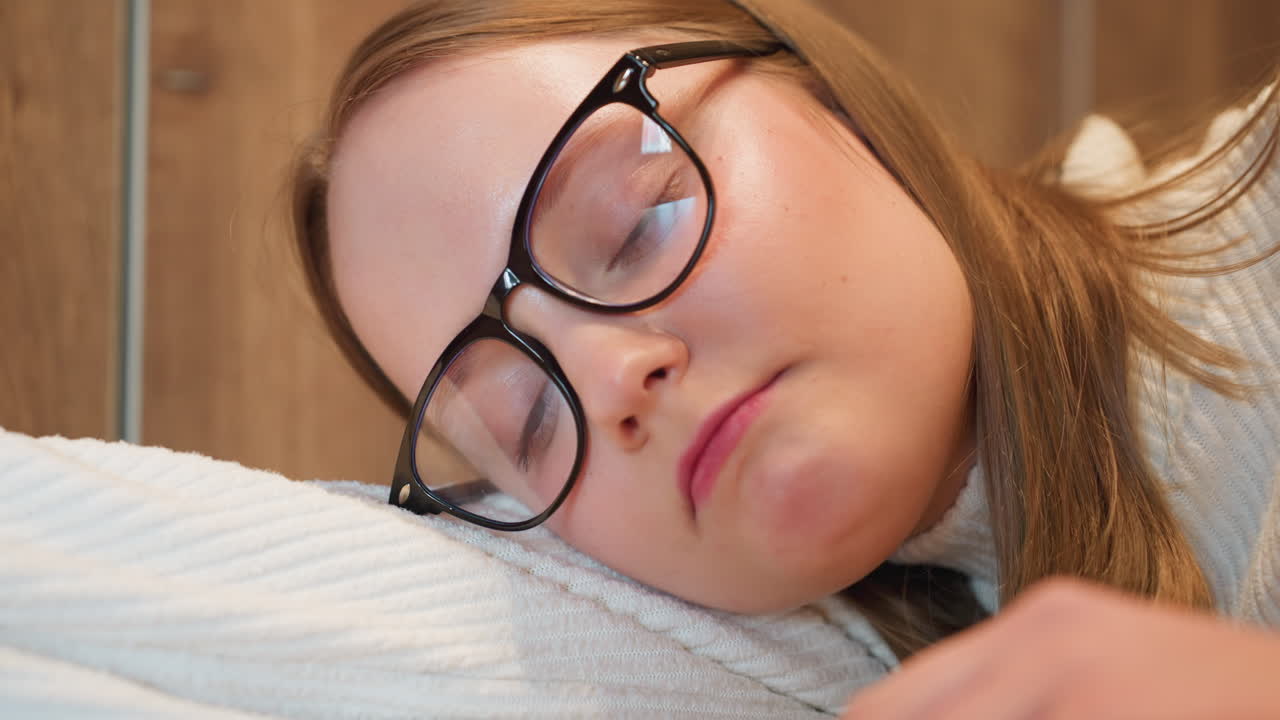 close up of young woman in glasses resting head on folded arms over wooden table, gazing quietly with thoughtful expression in cozy setting, wearing white ribbed sweater and wristwatch