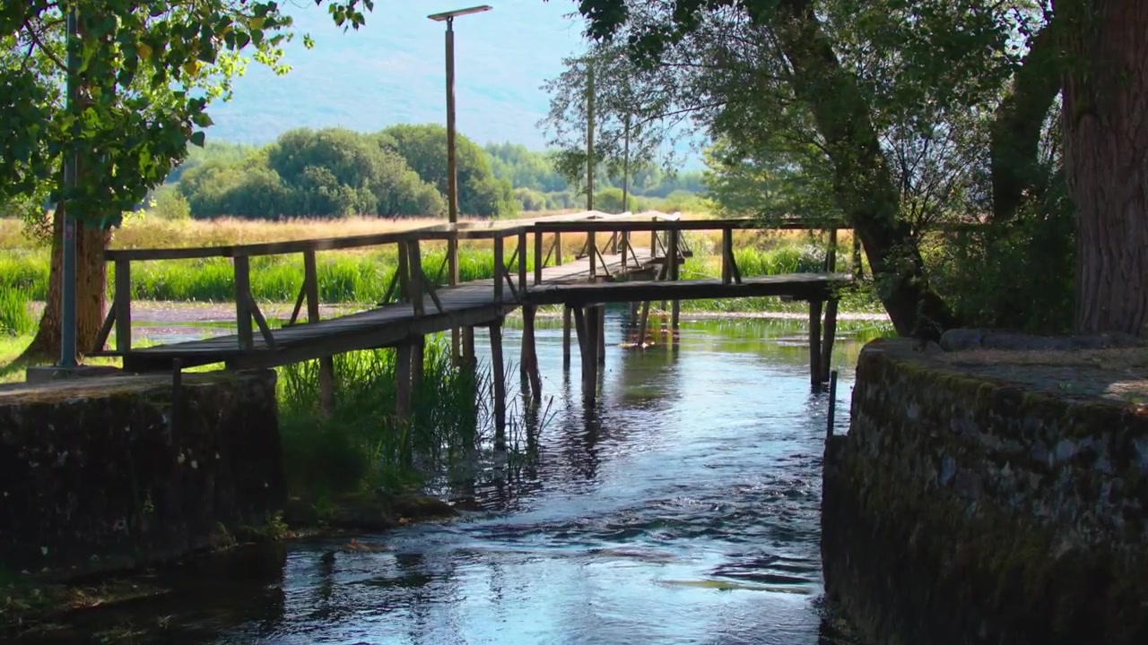 Passerella in legno che attraversa il limpido fiume Gacka vicino all'Hotel Gacka in Croazia, circondata da una vegetazione rigogliosa, alberi e un tranquillo paesaggio rurale