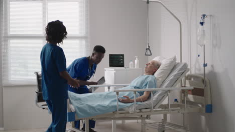 african young doctor team consulting covid-19 infected patient in bed at hospital. sick man lying in hospital bed and talking to surgeon in scrubs and protective mask. doctor examining elderly patient