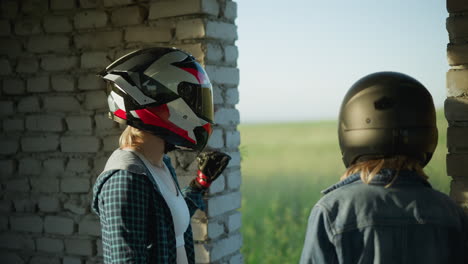 two women in helmets stand inside an abandoned building, one placing her hand on the wall while looking outside at the greenery