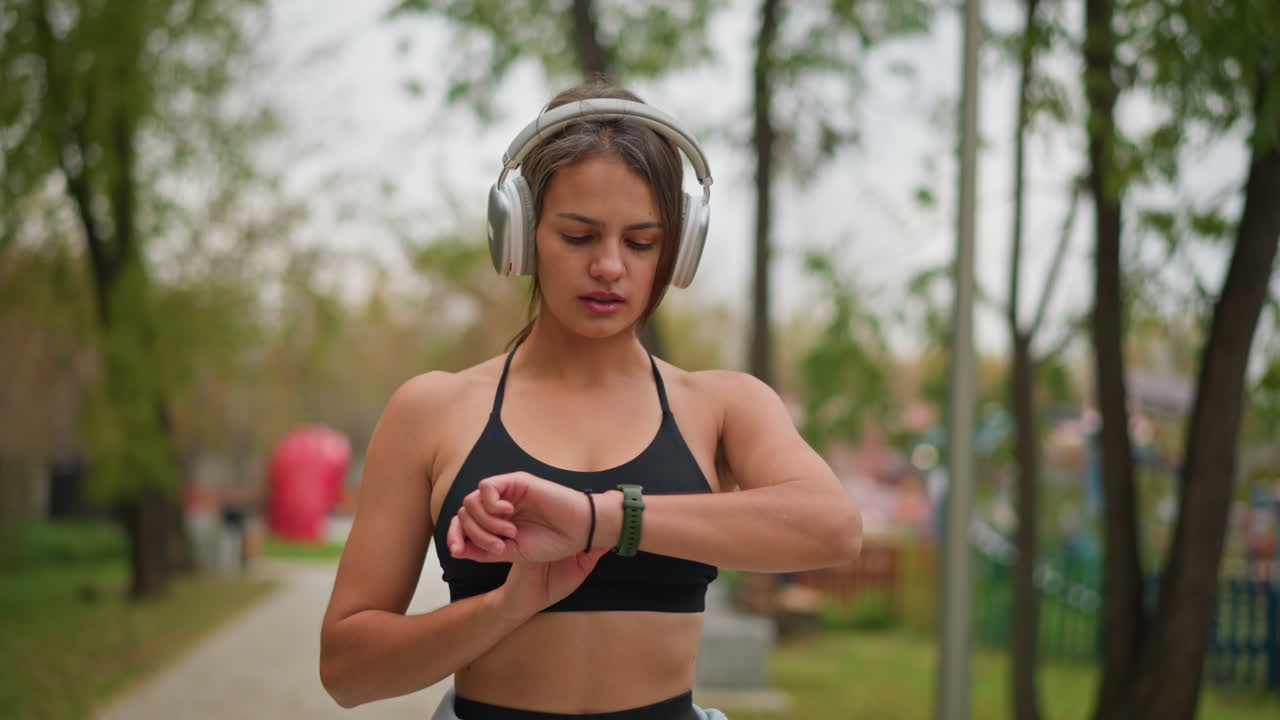 Woman in black singlet and headphones checking time on wristwatch in park, surrounded by trees, maintaining focus during workout session while keeping track of time for her exercise routine
