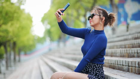 Frau-Im-Blauen-Pullover-Und-Sonnenbrille-Macht-Selfie,-Während-Sie-Auf-Außentreppen-Im-Stadtpark-Sitzt