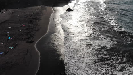 tropical beach aerial bird eye view of volcanic black sand and foaming ocean waves, people relaxing and swimming. bollullo beach, tenerife