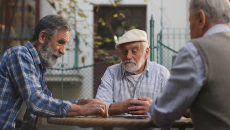 Un-Anciano-Feliz-Con-Una-Gorra-Ganando-Un-Juego-De-Dominó-De-Sus-Amigos-Vecinos-En-El-Patio-De-La-Mesa