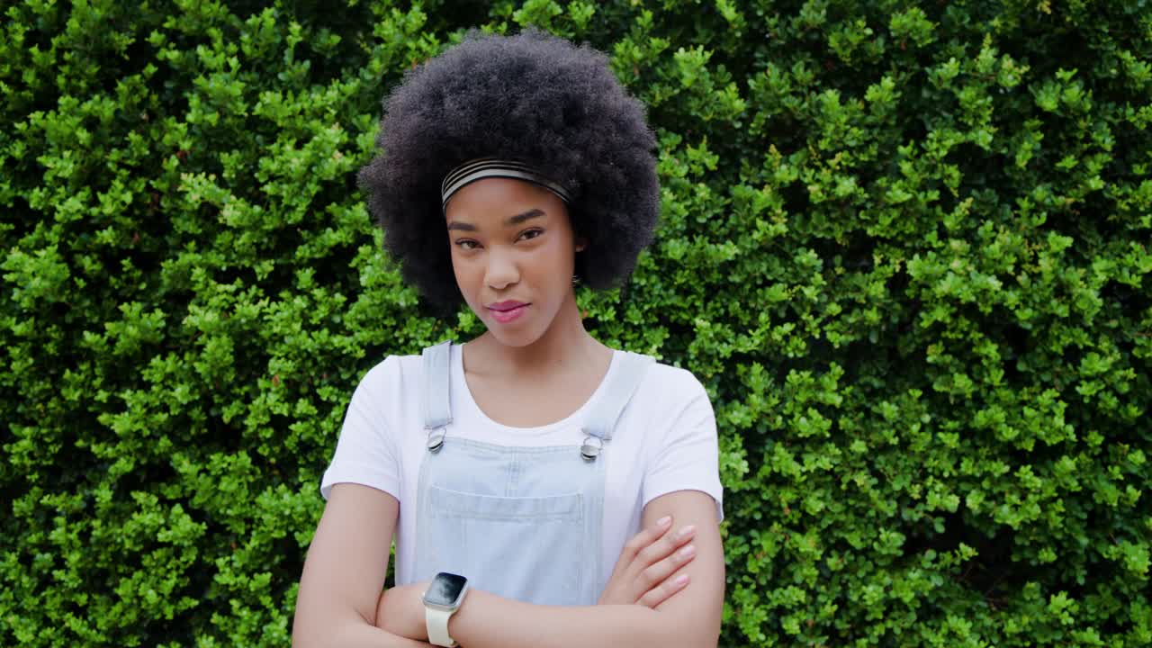 Afrikansk-amerikansk kvinde med krydsede arme foran en hæk, smilende med hårbånd, overalls, smartwatch.