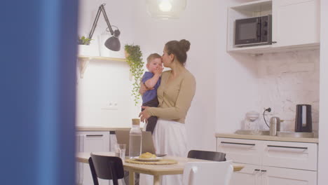 Young-Mother-Standing-And-Holding-Her-Baby-Boy-While-Working-With-Laptop-Computer-At-Home