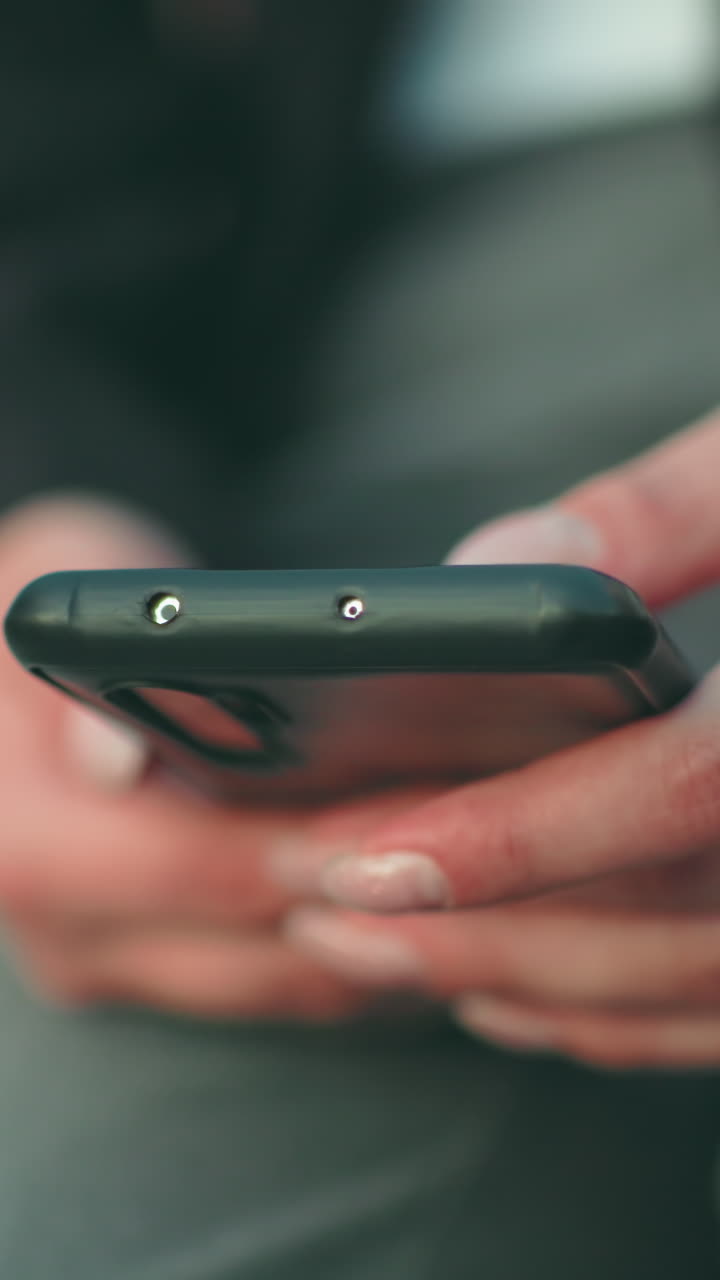 Close up soft focus of young lady with artificial nails operating phone in both hands while seated, wearing black jacket and wristwatch, with soft bokeh lights and blurred background
