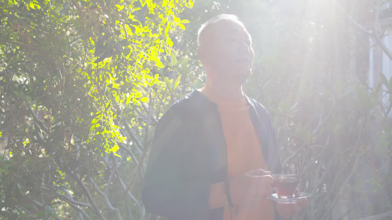 木漏れ日が差す庭で、ガラスのティーカップを持ち手すりのそばで飲んでいるアジア系の高齢男性