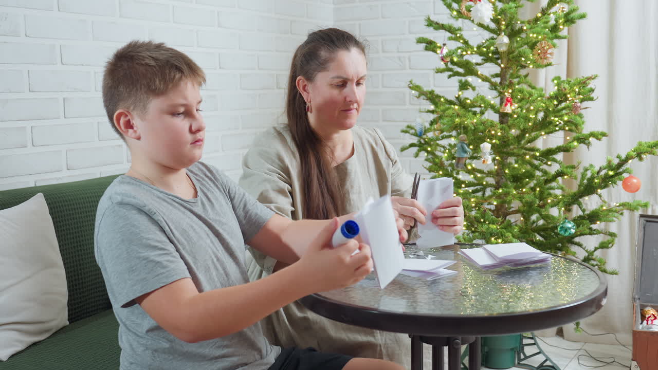 woman in beige gown and boy in grey shirt sitting at round table creating paper crafts using glue stick and pen with decorated christmas tree in background and soft white brick wall