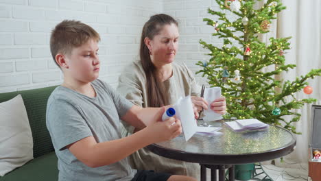 woman in beige gown and boy in grey shirt sitting at round table creating paper crafts using glue stick and pen with decorated christmas tree in background and soft white brick wall