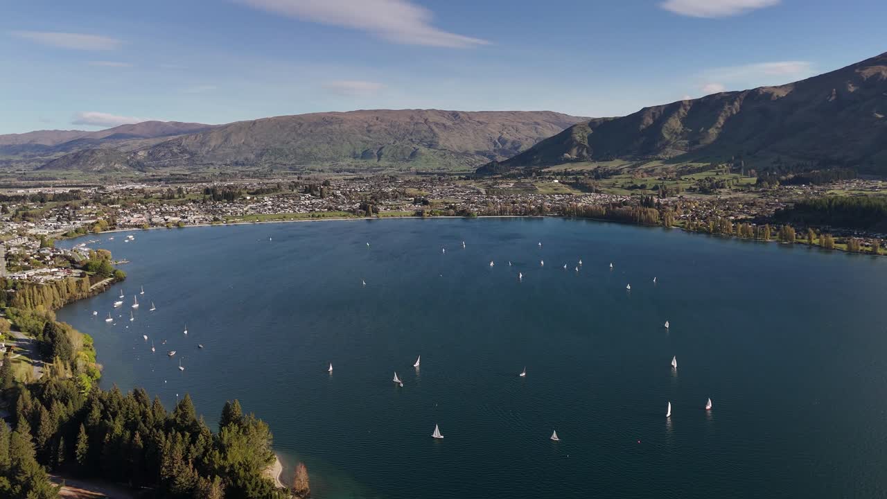 Una ripresa aerea cinematografica in 4K a 60fps con drone in retromarcia che rivela una flotta di barche a vela durante una regata sulle acque blu del Lago Wanaka, con le Alpi meridionali in lontananza, Nuova Zelanda.