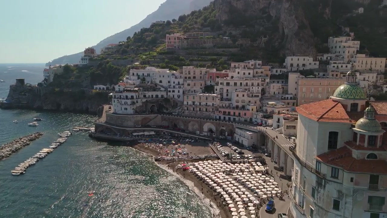 La Costiera Amalfitana con barche in acqua. Persone si rilassano in spiaggia sotto gli ombrelloni. Edifici colorati fiancheggiano le colline sopra il mare.