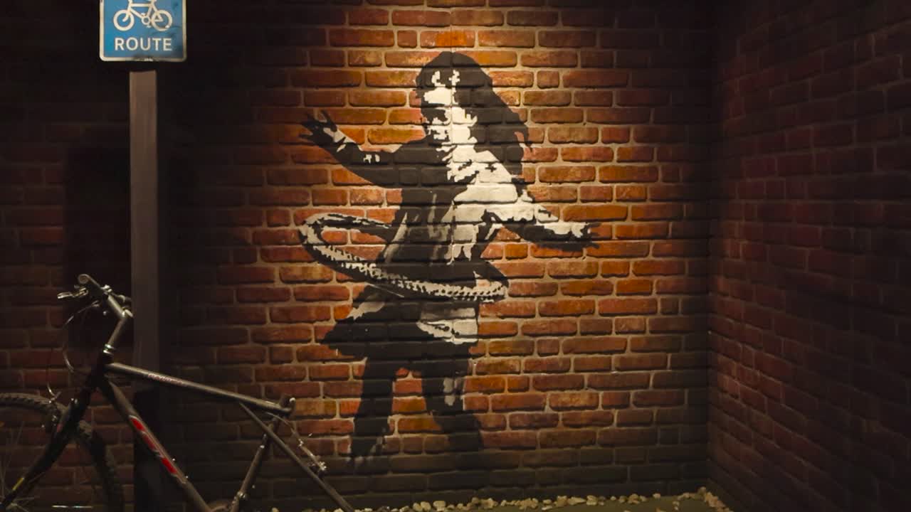 Stencil art or graffiti made by Banksy at his exhibition in Tallinn showing a small girl hula hooping with a bicycle tire done on a red and orange brick wall. Vintage bicycle in the foreground.