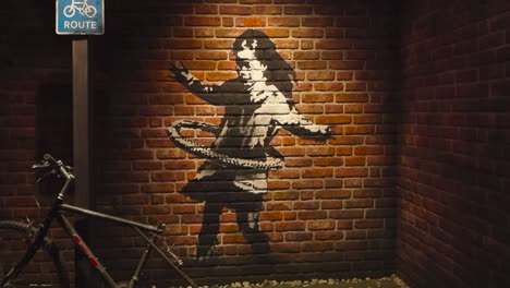 stencil art or graffiti made by banksy at his exhibition in tallinn showing a small girl hula hooping with a bicycle tire done on a red and orange brick wall. vintage bicycle in the foreground.