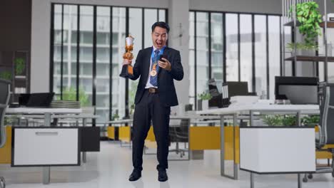 full body of asian business man in a suit and tie with a gold medal and trophy looking at smartphone then screaming goal celebrating in office
