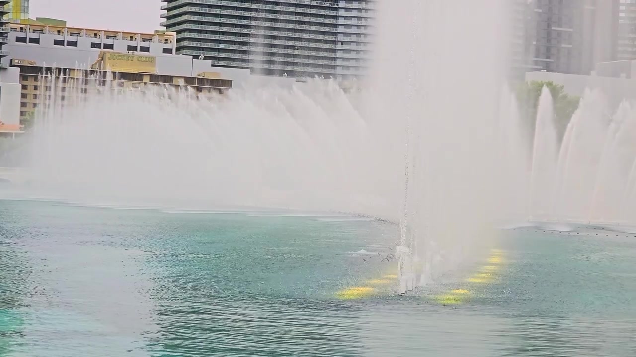 Toma panorámica del espectáculo de fuentes de Las Vegas con chorros de agua altos, lago turquesa, torres de hoteles, árboles, elegantes arcos de piedra, el horizonte del resort y un ambiente turístico soleado.
