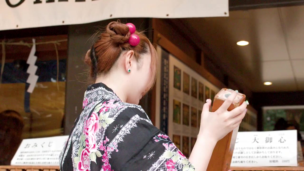A woman wearing a traditional floral kimono shakes a wooden fortune box at a shrine