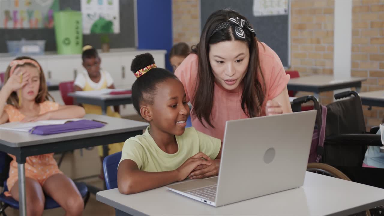 Happy diverse female teacher helping girl using laptop in elementary school class, slow motion