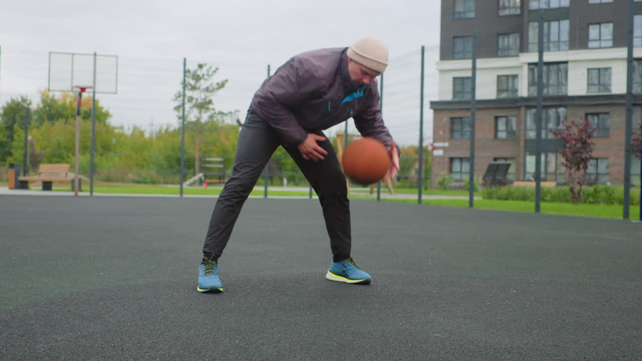 man dribbling basketball on court, residential backdrop with apartment buildings, overcast sky, low camera angle highlighting asphalt texture and sneakers, focused solo workout with footwork drills