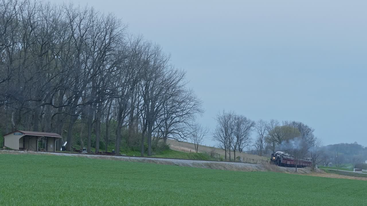 A vintage steam train chugs along a rural track, emitting dark smoke as it moves past a small shelter surrounded by trees. The tranquil landscape reflects evening ambiance.