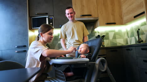 caucasian woman is feeding her lovely baby. mother tries the food from the spoon. man stands behind smiling. low angle view.