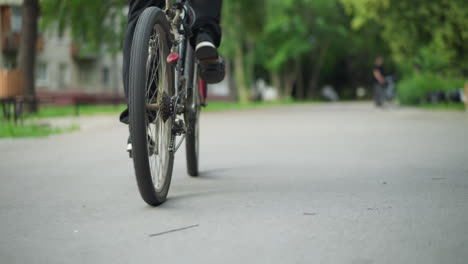 vista trasera de alguien con zapatillas negras y pantalones negros, pedaleando bicicleta lentamente a lo largo de un camino pavimentado, la escena presenta un fondo borroso con alguien de pie y vegetación