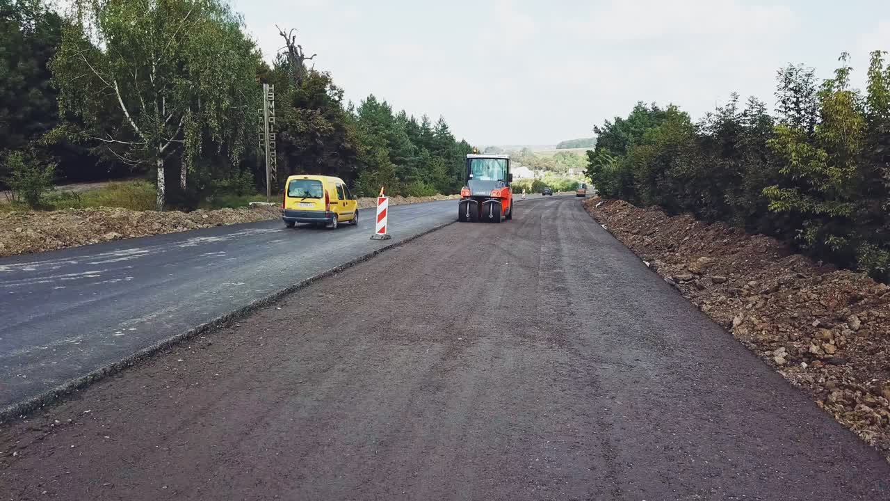 A modern vibratory roller of red color tampers with fresh asphalt on the road on the background of green trees and traffic outside the city. Aerial view.