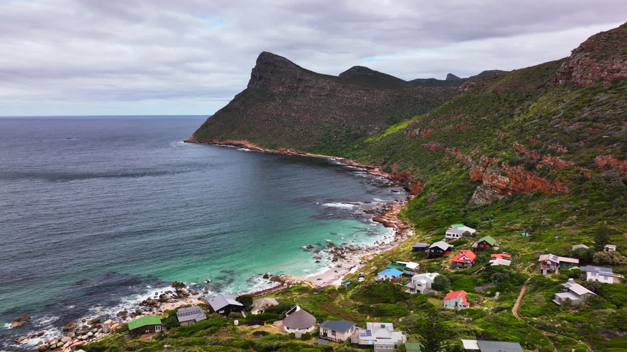 South Africa small beach town Smitswinkel Bay Cape of Good Hope Simon's Town aerial drone cinematic homes surf wave green lush spring summer deep aqua blue sea wind cloudy mountain backward motion