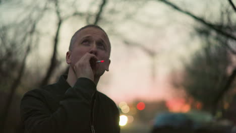 a man in a green jacket smoking a cigarette outdoors at dusk, the background is blurred with soft, warm lights