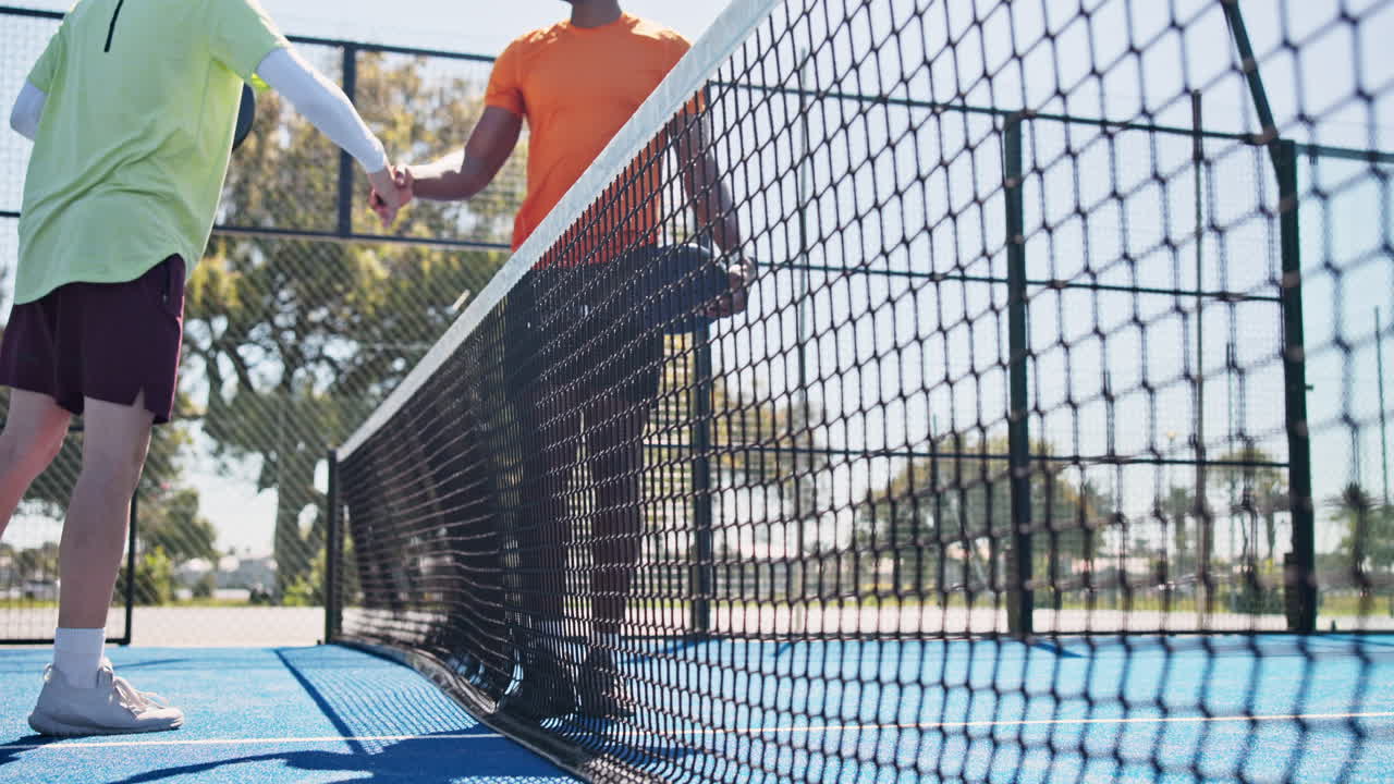 Zwei Männer schütteln sich über einem Padel-Tennisnetz auf einem sonnigen Platz die Hände.