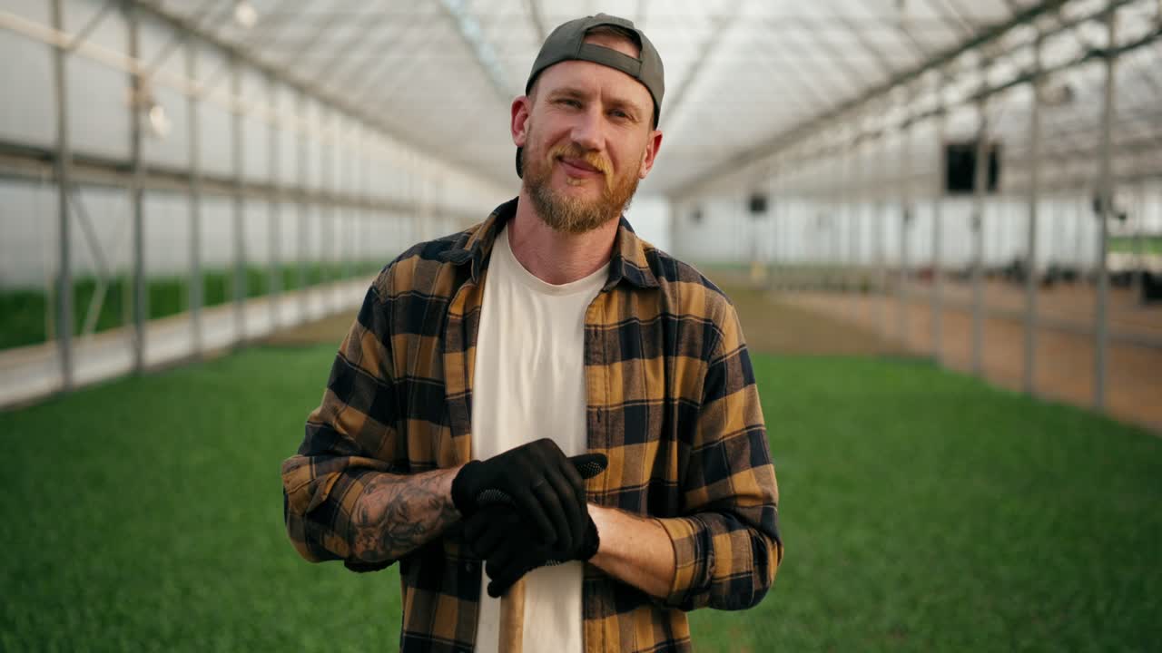retrato de un hombre agricultor confiado en una gorra con una barba en una camisa a cuadros que posa entre los brotes de plantas jóvenes en un invernadero en la granja