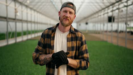 Portrait-of-a-confident-farmer-guy-in-a-cap-with-a-beard-in-a-plaid-shirt-who-poses-among-the-sprouts-of-young-plants-in-a-greenhouse-on-the-farm