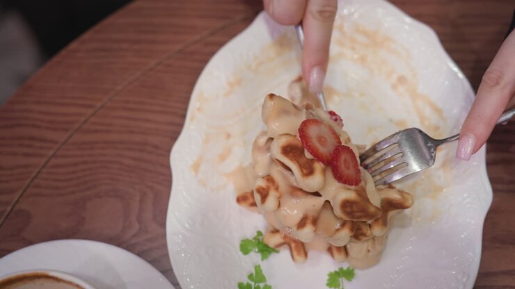 Close up of lady using fork and knife to cut through creamy waffles topped with fresh strawberries and garnished with green leaves, placed on white textured plate on wooden table beside coffee cup