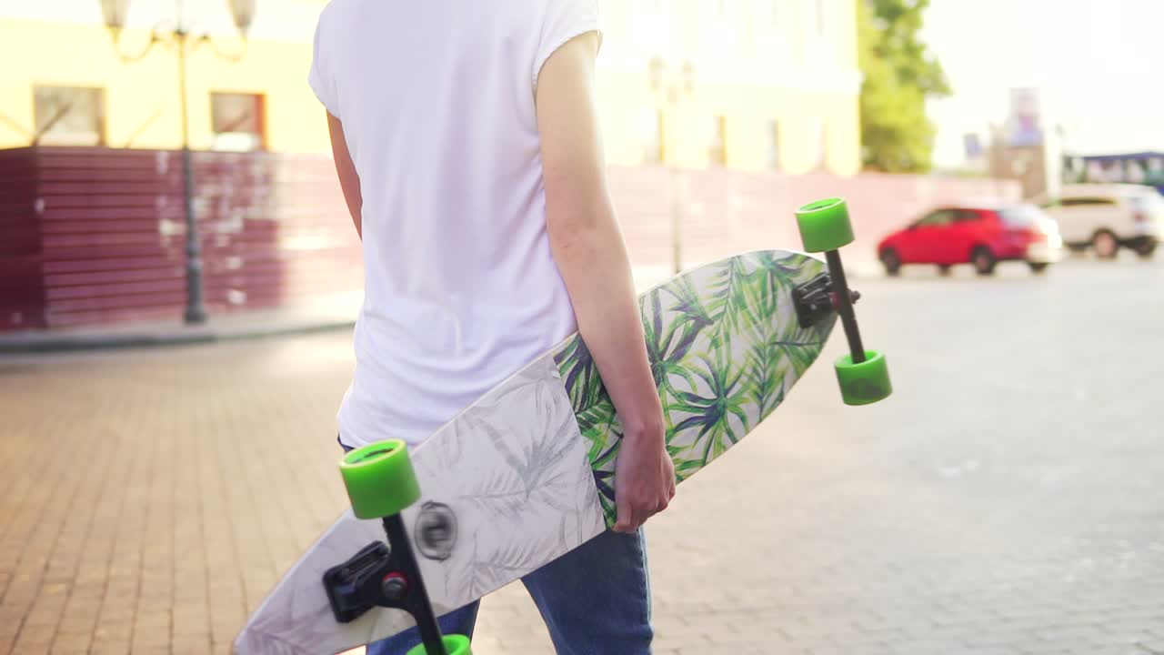 Back view of a woman walking in the old city street holding her trendy stylish longboard with flowers in the morning. Lens flare