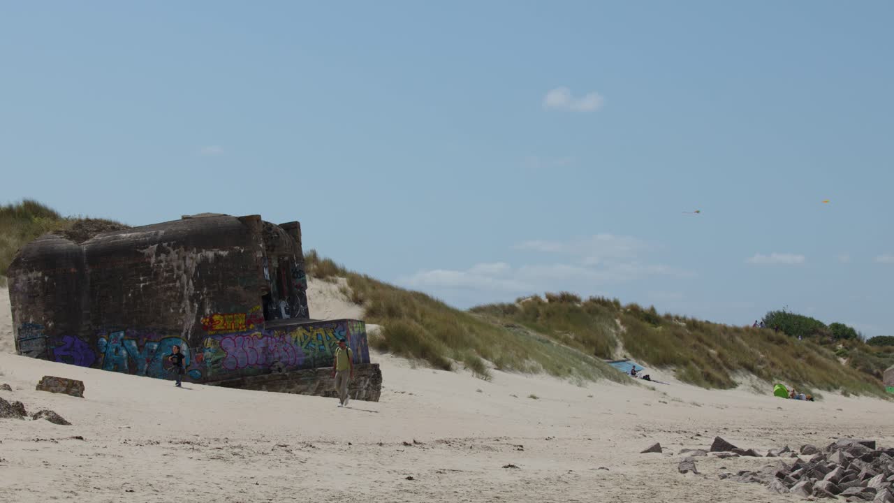 Family strolls by colorful graffiti bunker on sandy beach, bright daylight, static wide shot