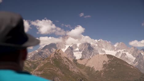hombre con un sombrero mirando un gran pico montañoso cubierto de nubes en un día soleado en los alpes