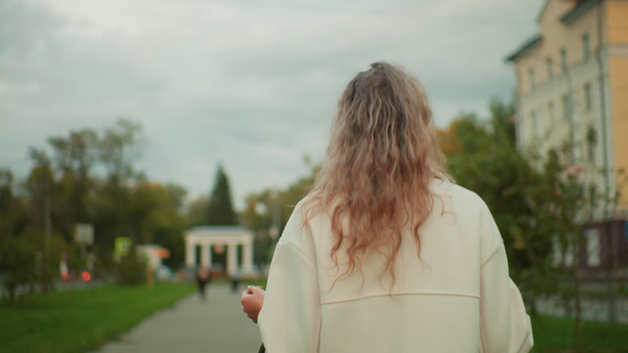 White girl walking down city sidewalk twirling closed umbrella while wearing light jacket, back view, relaxed mood, green park surroundings, soft natural lighting, cloudy sky, urban life atmosphere