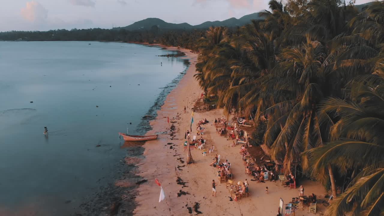 воздушный полет над людьми, сидящими у бара на пляже тропического острова во время заката