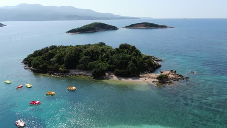 Drone-view-in-Albania-flying-in-front-of-three-green-small-islands,-blue-crystal-clear-water-and-pedal-color-boats,-sunny-day-in-Ksamil