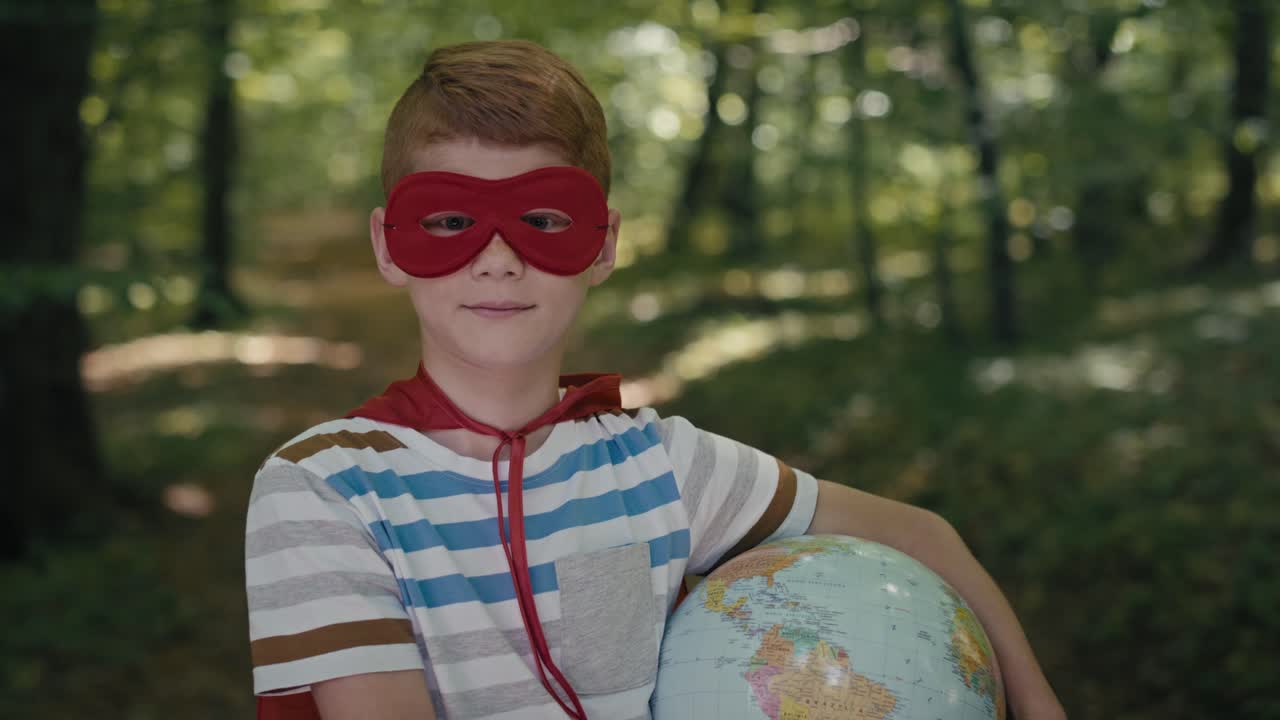 슈퍼히어로 의상을 입은 홍색 소년의 초상화가 숲에서 지구를 들고 있습니다.