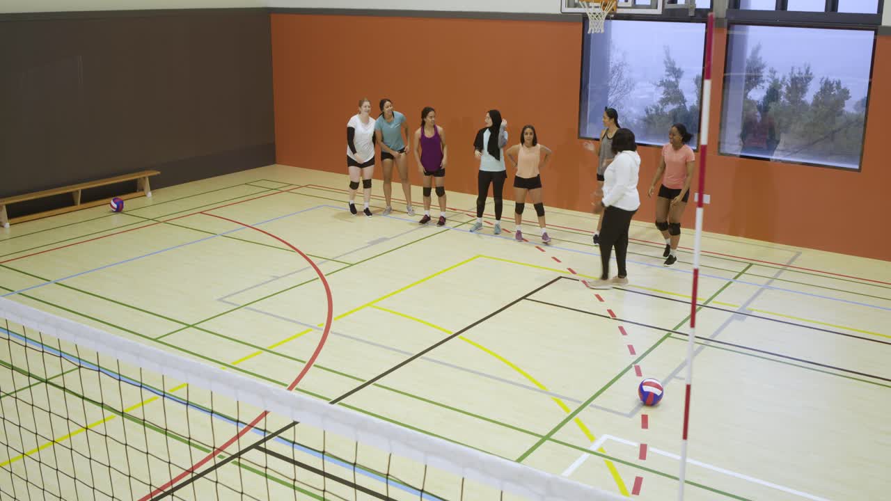 Et mangfoldigt kvindevolleyboldhold bevæger sig langs nettet i en skolehal, efter at træneren har signaleret start på øvelsen.