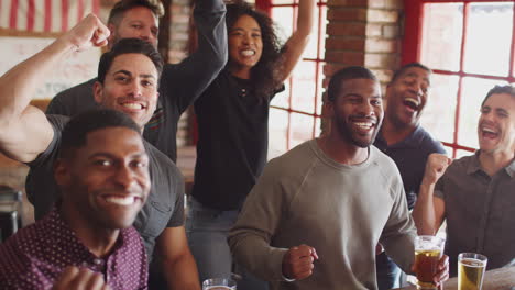 group of male and female friends celebrating whilst watching game on screen in sports bar