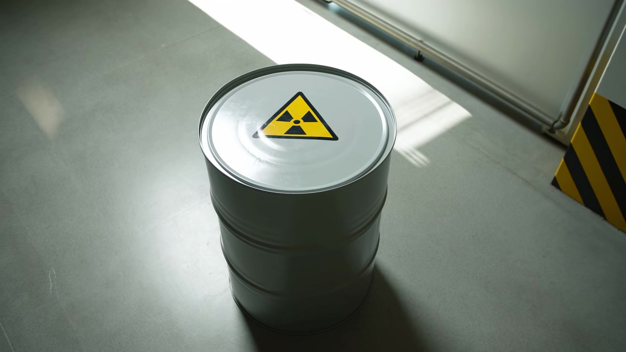 Gray steel barrel with clear radiation hazard emblem stays in place while camera subtly moves, revealing hard floor reflections, strong side light, and striped industrial barrier.