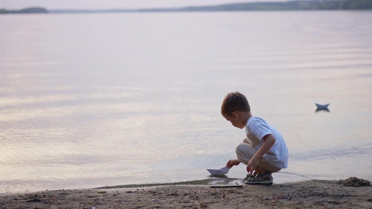Anak laki-laki berjongkok meletakkan perahu kertas di atas air danau yang tenang. Tampak samping anak berjongkok di tepi berpasir meluncurkan perahu origami saat senja.