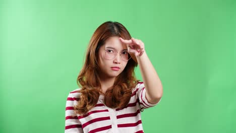 young asian woman with eyeglasses pointing to the eyes watching you gesture, keeping eyes on you, green screen background