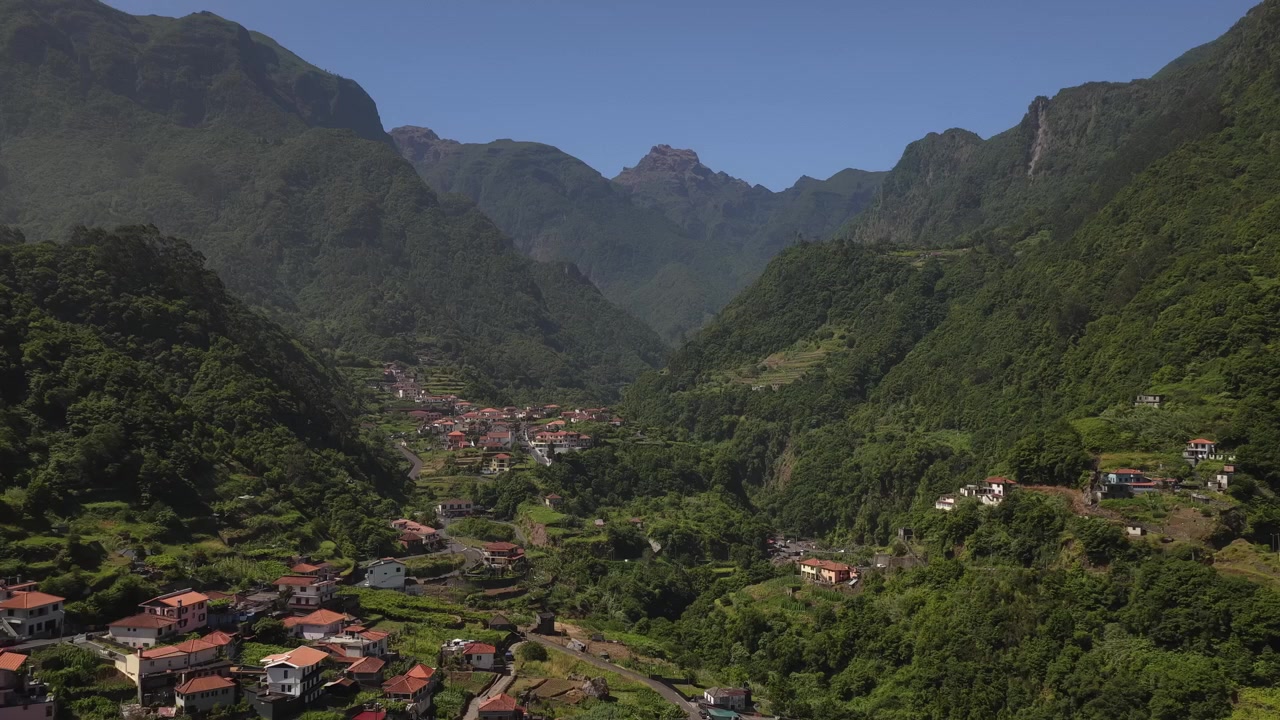 Riprese aeree sorvolano Faja do Penedo, rivelando case dai tetti rossi immerse in una lussureggiante valle di foresta di lauro protetta dall'UNESCO, sotto imponenti cime montuose e cieli azzurri e sereni.