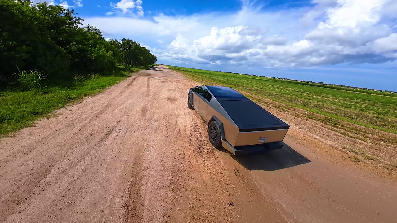 Flight above the agricultural field approaching a technological Tesla Cybertruck riding by the ground road. Futuristic auto avoiding puddles on the track. FPV drone footage.